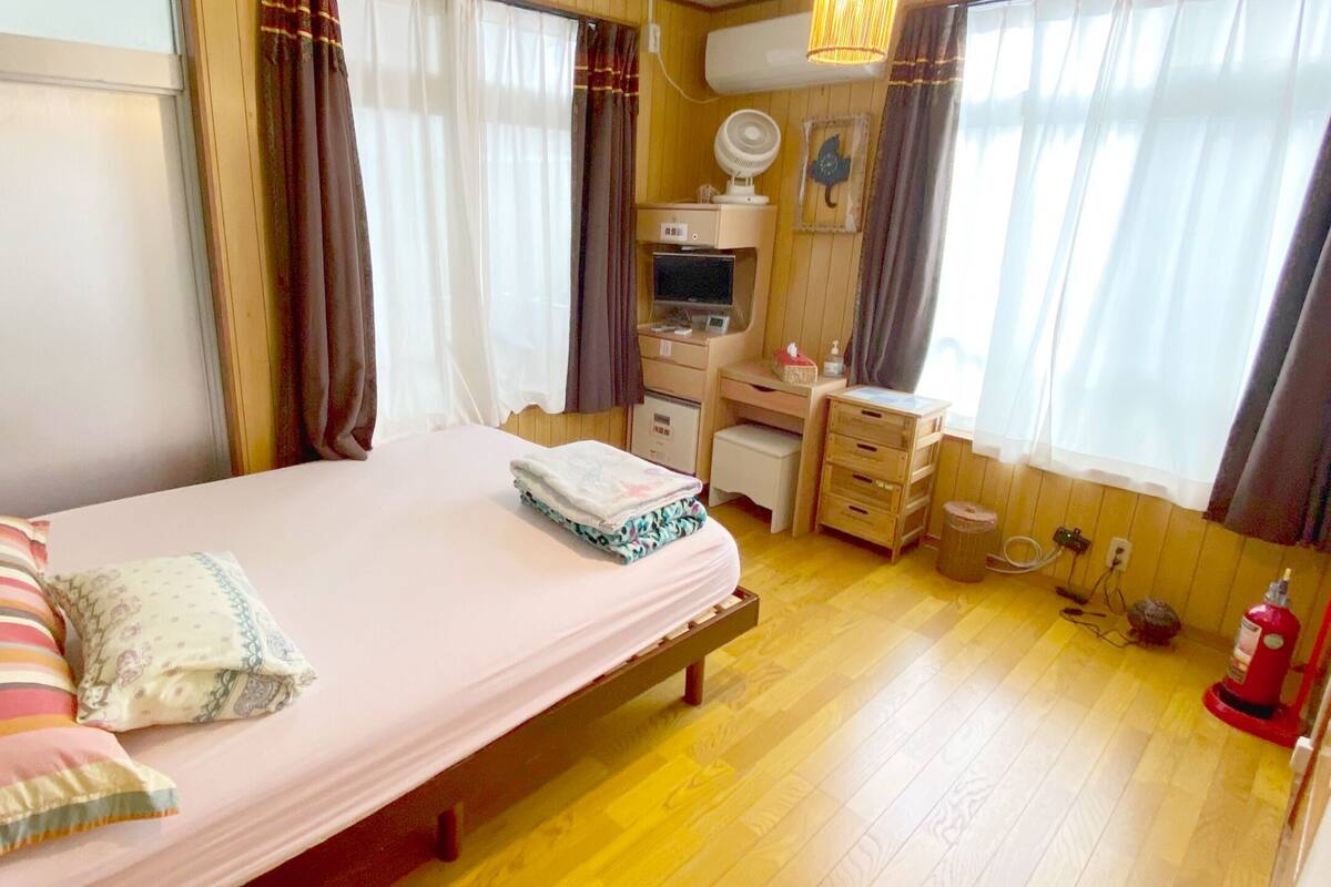 Un dormitorio con cama, mesita de noche, escritorio, silla y ventana con cortinas.