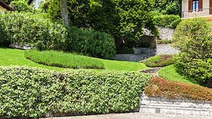 A landscaped garden with a stone wall, a curved pathway, and a building with a brown roof and windows. - Cernobbio Lake View (Cernobbio)
