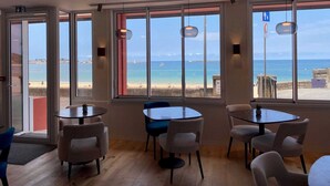 Een kamer met grote ramen met uitzicht op een strand, met verschillende ronde tafels en stoelen.