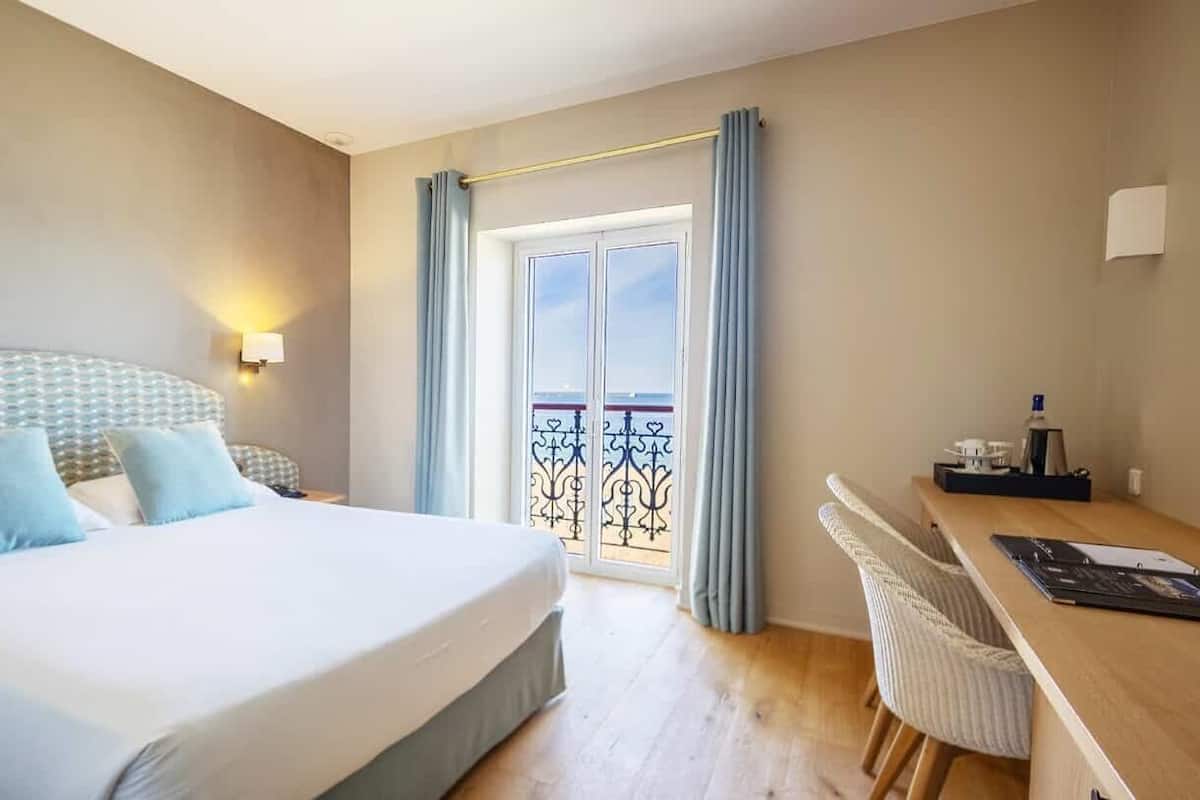 A hotel room with a bed, a desk, a chair, a TV, and a balcony with curtains.