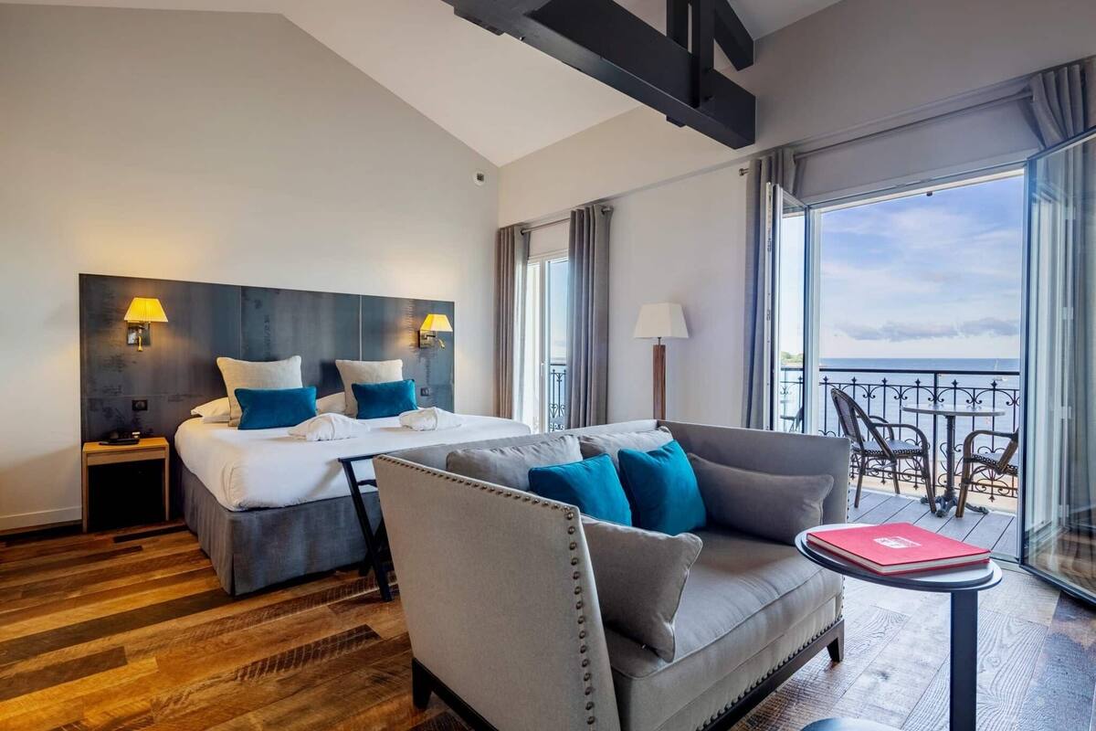 A hotel room with a bed, a sofa, a small table, and a balcony with a view.