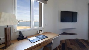 Een kamer met een houten bureau, een lamp, een aan de muur bevestigde televisie en uitzicht op zee door een raam.