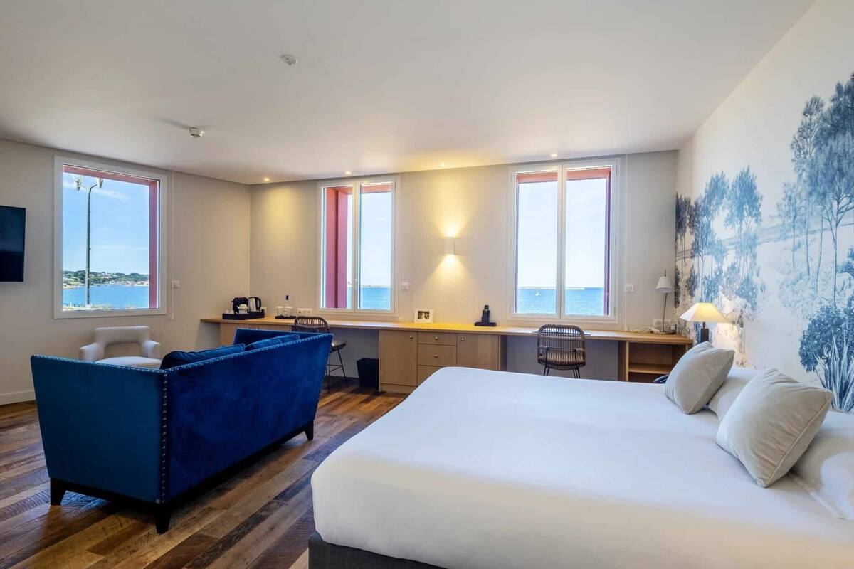 A hotel room with a bed, a blue sofa, a desk, and a view of the sea.
