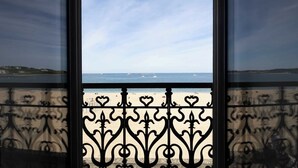 Vista a través de una ventana con barandillas de hierro decorativas, que da a una playa con barcos y un cielo despejado.