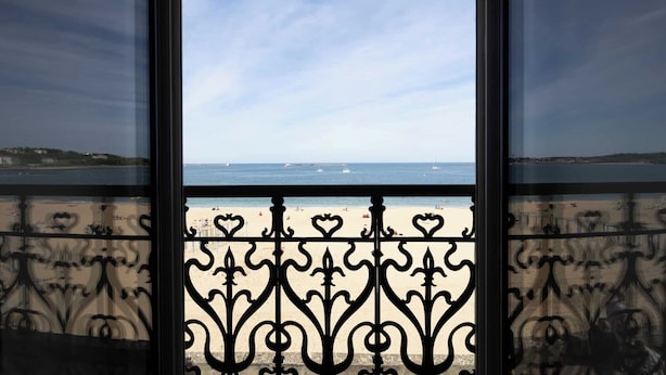 Uitzicht door een raam met sierijzeren hekwerk, op een strand met boten en een heldere lucht.
