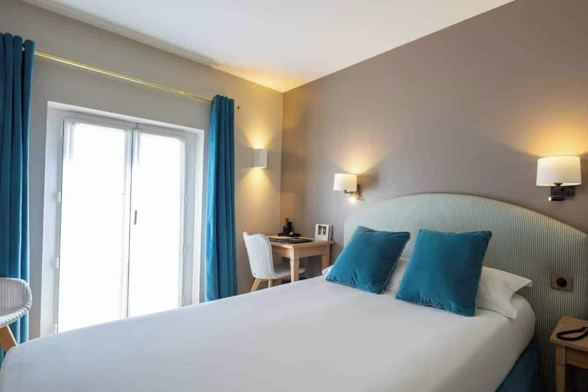 A hotel room with a bed, a desk, a chair, a TV, and two wall-mounted lamps.