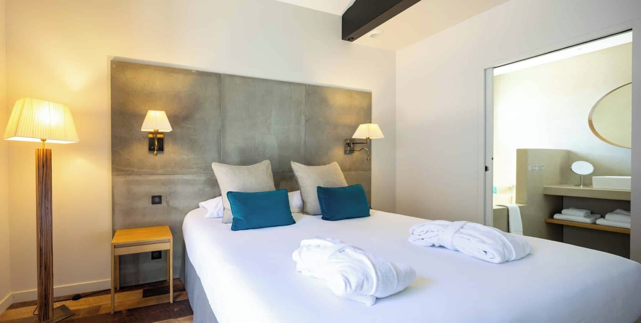 Ein Hotelzimmer mit einem großen Bett, zwei Wandlampen, einem Nachttisch, einem kleinen Hocker und einem Badezimmer mit Spiegel und Ablagen.