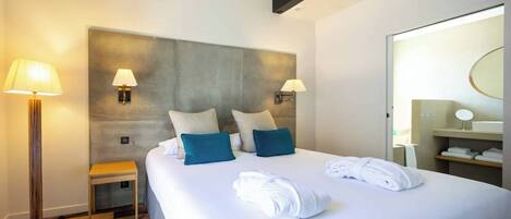 Ein Hotelzimmer mit einem großen Bett, zwei Wandlampen, einem Nachttisch, einem kleinen Hocker und einem Badezimmer mit Spiegel und Ablagen.