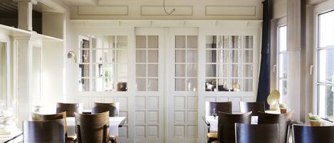 A dining area with wooden tables and chairs, white doors with glass panels, and a view of the outdoors through large windows.