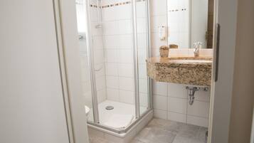 Un baño moderno con ducha de vidrio, lavamanos con encimera de granito y suelo de baldosas blancas.