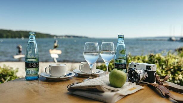 A table with a camera, two glasses of water, a bottle, a cup, a saucer, a spoon, a green apple, and a newspaper. - Hotel Wassersleben (Harrislee)