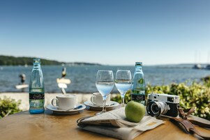 Una mesa con una cámara, dos vasos de agua, una botella, una taza, un platito, una cuchara, una manzana verde y un periódico.