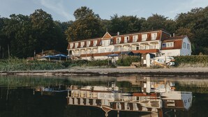 A waterfront hotel with multiple balconies overlooking the water, a dock with boats, and a reflection in the calm water. - Hotel Wassersleben (Harrislee)