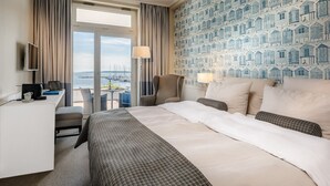 A hotel room with a bed, desk, chair, and a view of the water with boats. - Hotel Wassersleben (Harrislee)