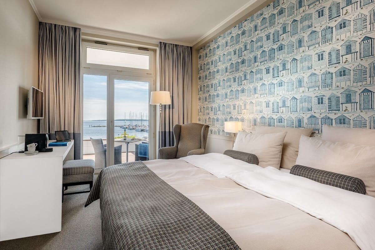 A hotel room with a bed, desk, chair, and a view of the water with boats.
