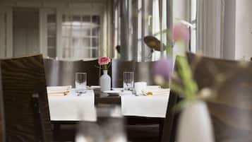 Una mesa de comedor dispuesta con manteles blancos, platos, vasos y cubiertos. Hay un florero con una flor rosa sobre la mesa.