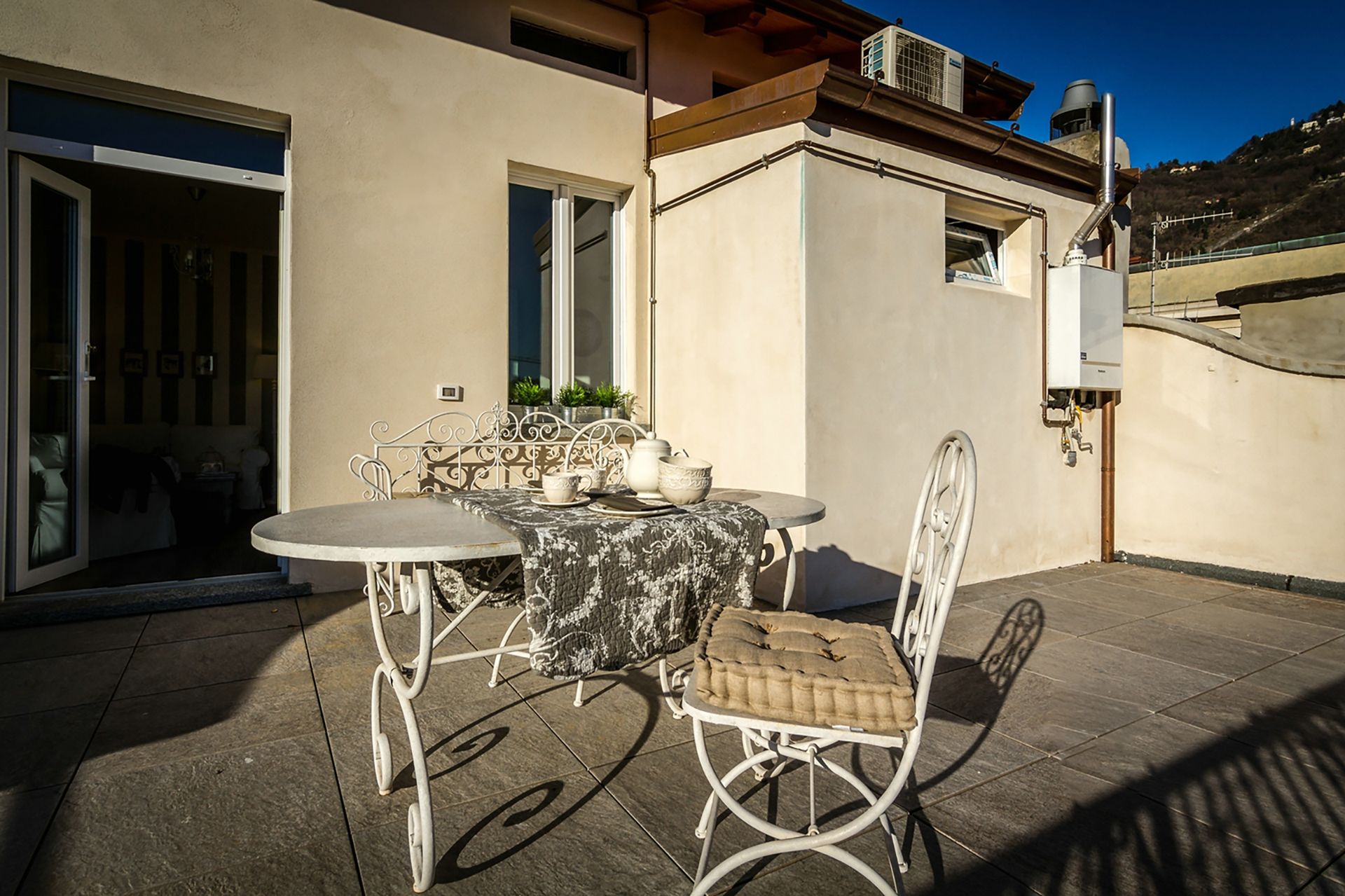Un patio exterior con una mesa redonda y dos sillas, una mesita con objetos decorativos, y un ventanal corredero que da a una habitación interior.
