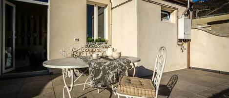 Un patio exterior con una mesa redonda y dos sillas, una mesita con objetos decorativos, y un ventanal corredero que da a una habitación interior.