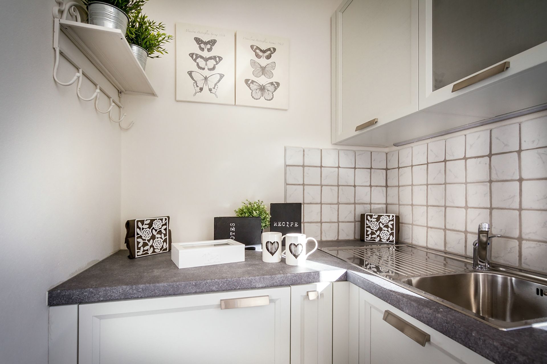 Een keuken met witte kastjes, een granieten aanrecht, een roestvrijstalen spoelbak en decoratieve elementen, waaronder een plant, een lijst met vlinderkunst en een receptbord.