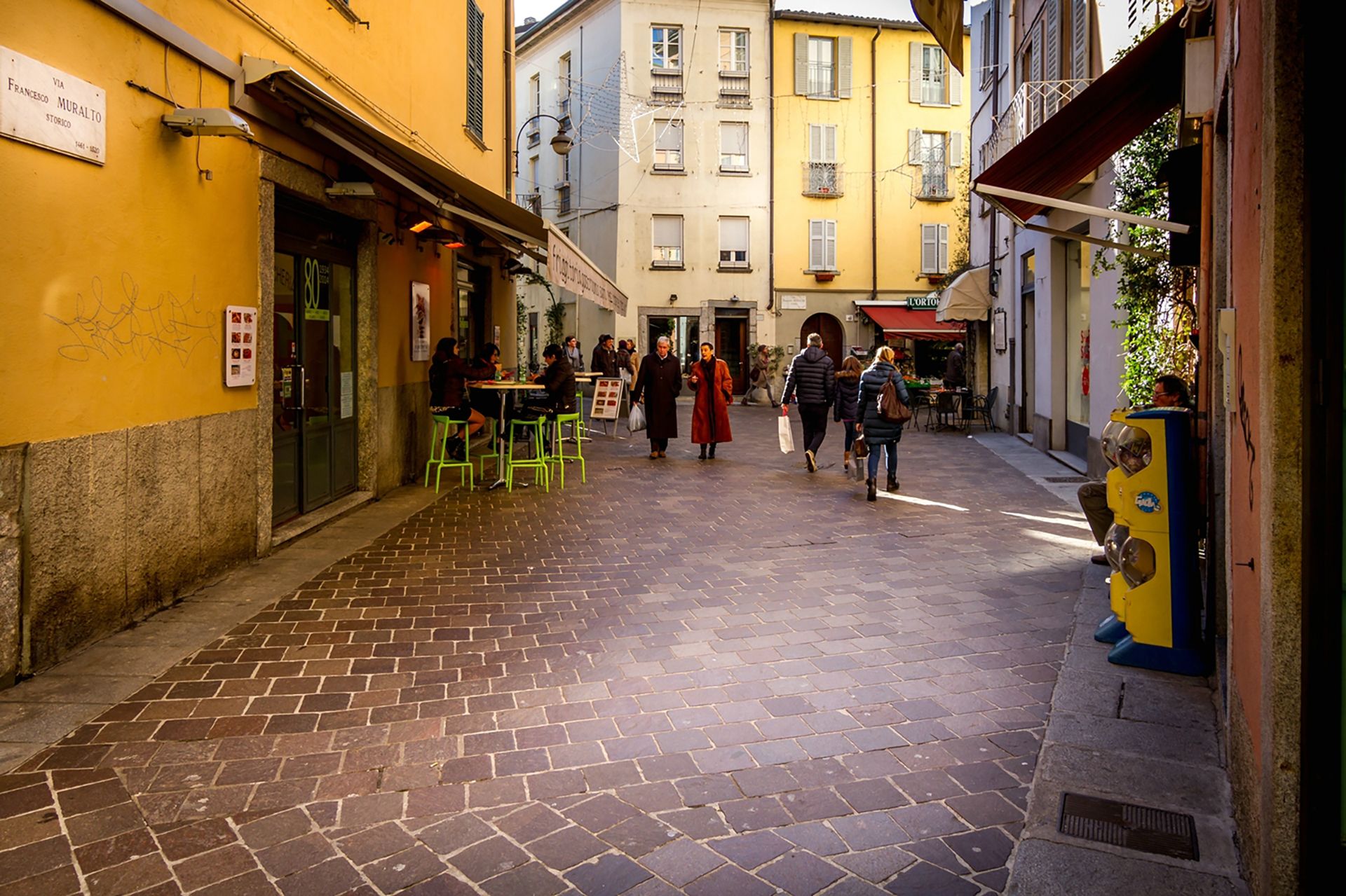 A cobblestone street with outdoor seating, buildings on either side, and a few pedestrians.