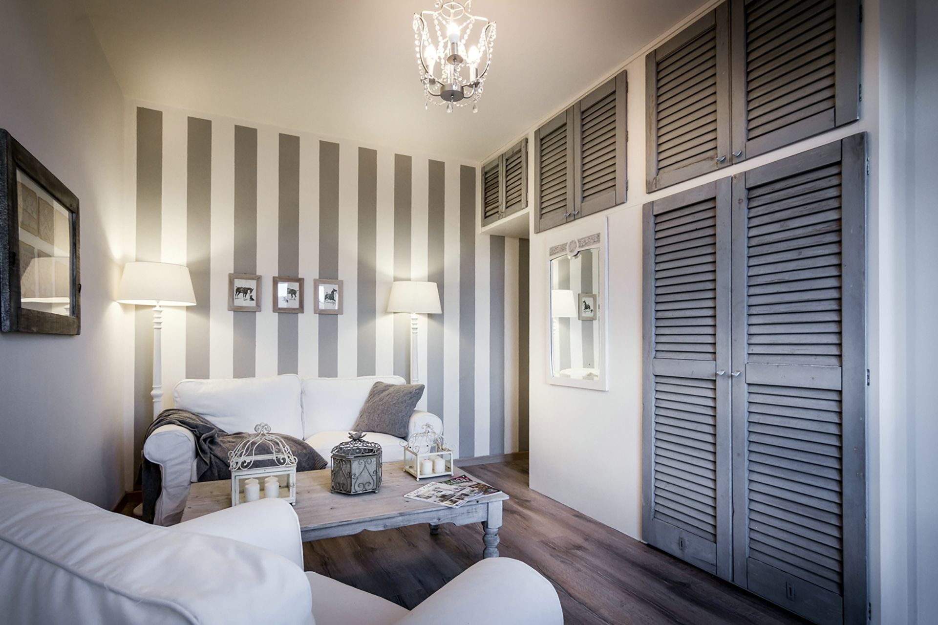 Een kamer met een gestreepte wand, een kroonluchter, een witte bank en een houten tafel met decoratieve objecten.