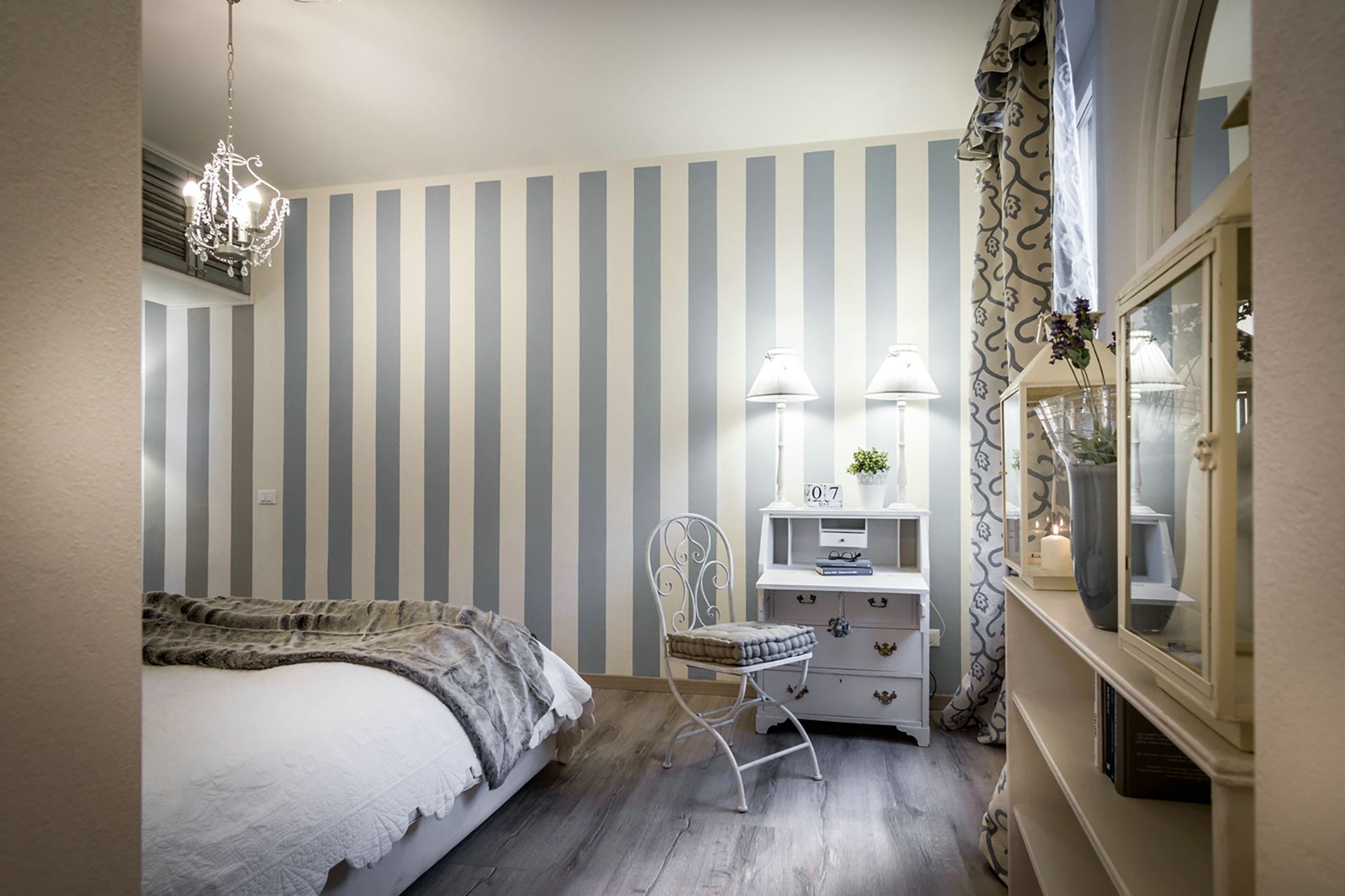 A bedroom with striped wallpaper, a bed with a grey blanket, a white chair, a nightstand with a lamp, and a dresser with a mirror.