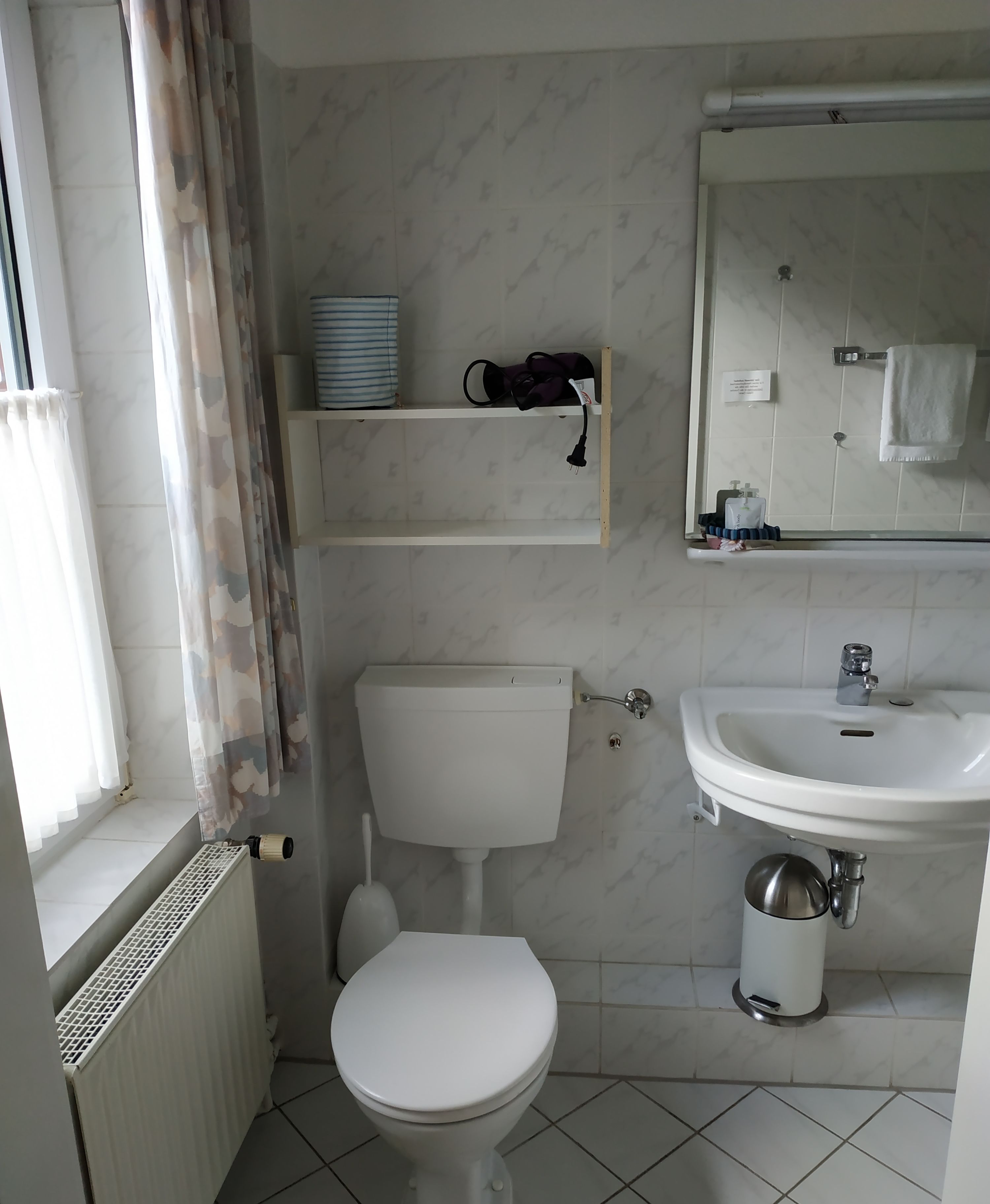 Ein Badezimmer mit Toilette, Waschbecken und Spiegel. Es gibt eine Heizung, ein Ablagefach mit Pflegeprodukten und ein Fenster mit Vorhängen.