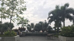 Een terras buiten met stenen vloer, zitgedeeltes en diverse planten, waaronder palmbomen en struiken.