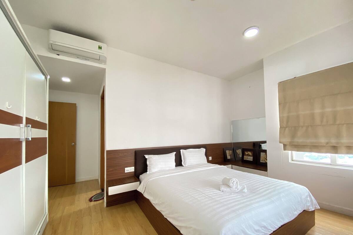 Een slaapkamer met een houten bed, wit beddengoed, een plafondairconditioner, een raam met een zonnescherm en een houten kledingkast.