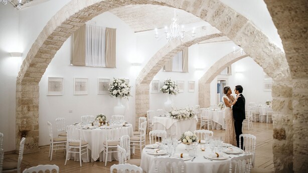 A wedding reception in a rustic venue with stone arches, white tables, and floral centerpieces.