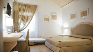 A hotel room with a bed, a desk, a chair, and a small table. There are curtains on the window, and the walls have framed pictures.