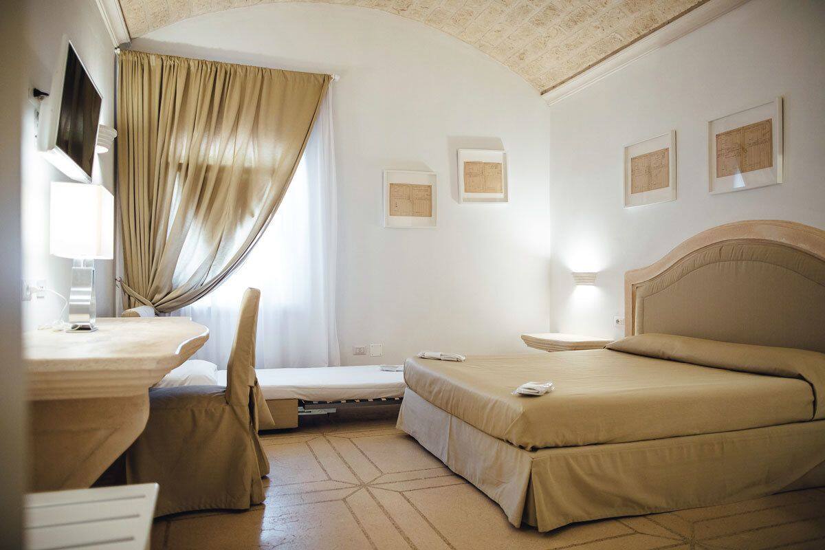 A hotel room with a bed, a desk, a chair, and a small table. There are curtains on the window, and the walls have framed pictures.