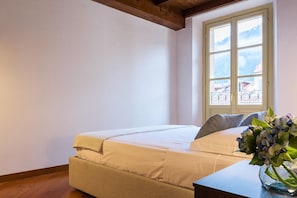 Quarto com cama, janela com vista para edifícios e um vaso com flores em uma mesa de cabeceira.