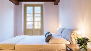 Une chambre à coucher avec un lit, une table de chevet avec une lampe, un vase de fleurs et une fenêtre donnant sur des immeubles.