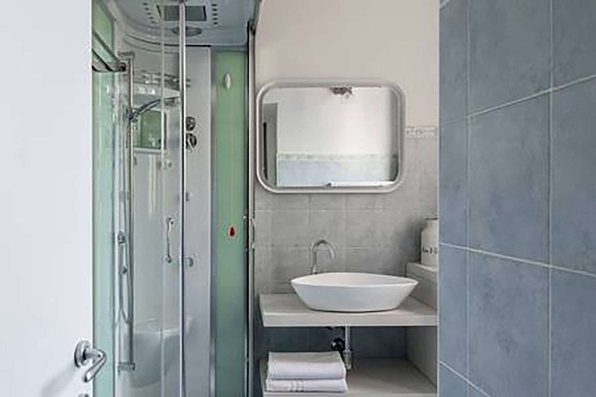 Un baño pequeño y funcional con cabina de ducha de vidrio, lavamanos blanco y un espejo reducido.