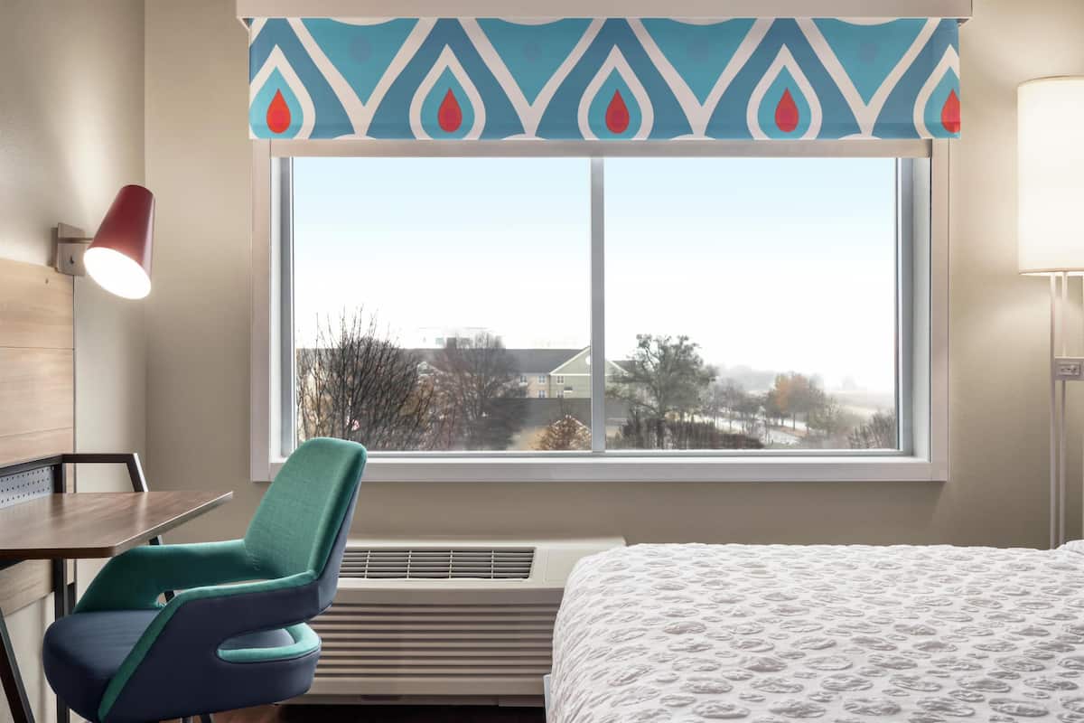 A hotel room with a bed, a desk, a chair, a window with a view, and a wall-mounted lamp.