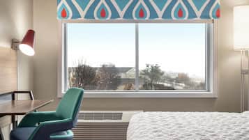 A hotel room with a bed, a desk, a chair, a window with a view, and a wall-mounted lamp.