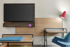 Una habitación con cama, escritorio, silla, televisor montado en la pared y una lámpara roja.