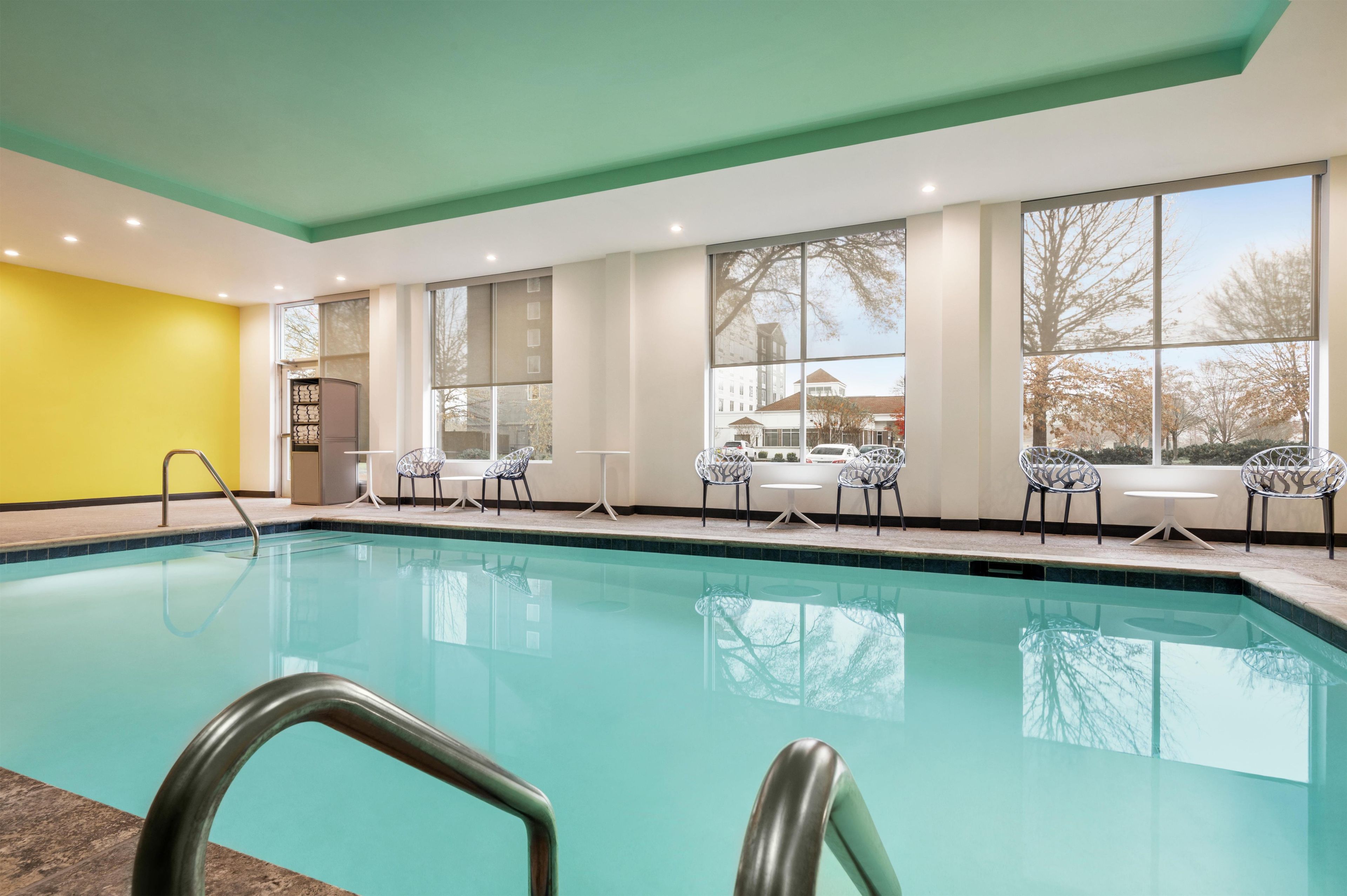 Une piscine intérieure avec des parois en verre transparent, un mur jaune d’un côté et une vue sur un bâtiment à l’extérieur.