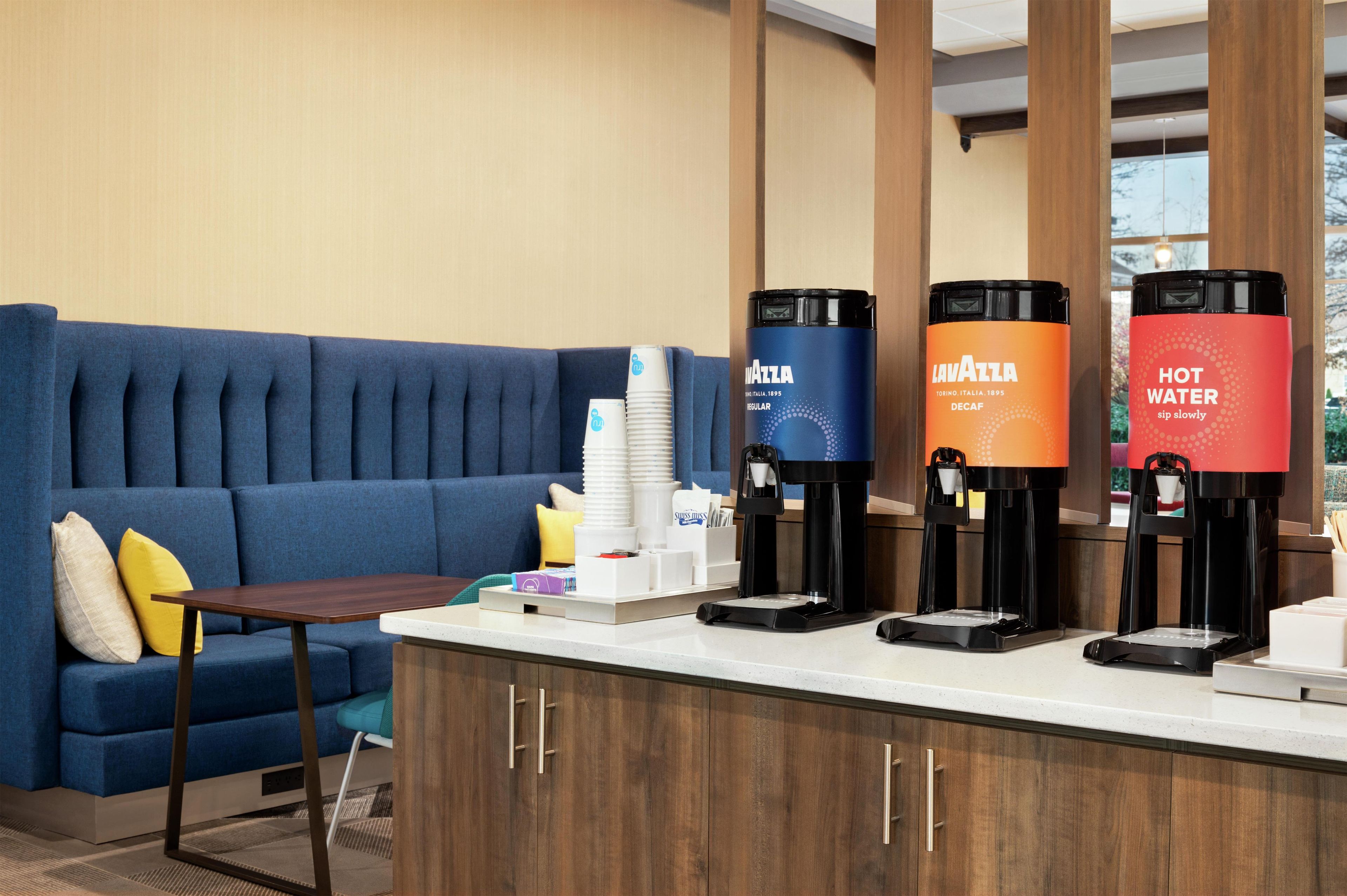 Een hotel receptie met een blauwe bank, een klein tafeltje en drie drank automaten, gelabeld met 'MALTA', 'MALTA' en 'HEET WATER'.