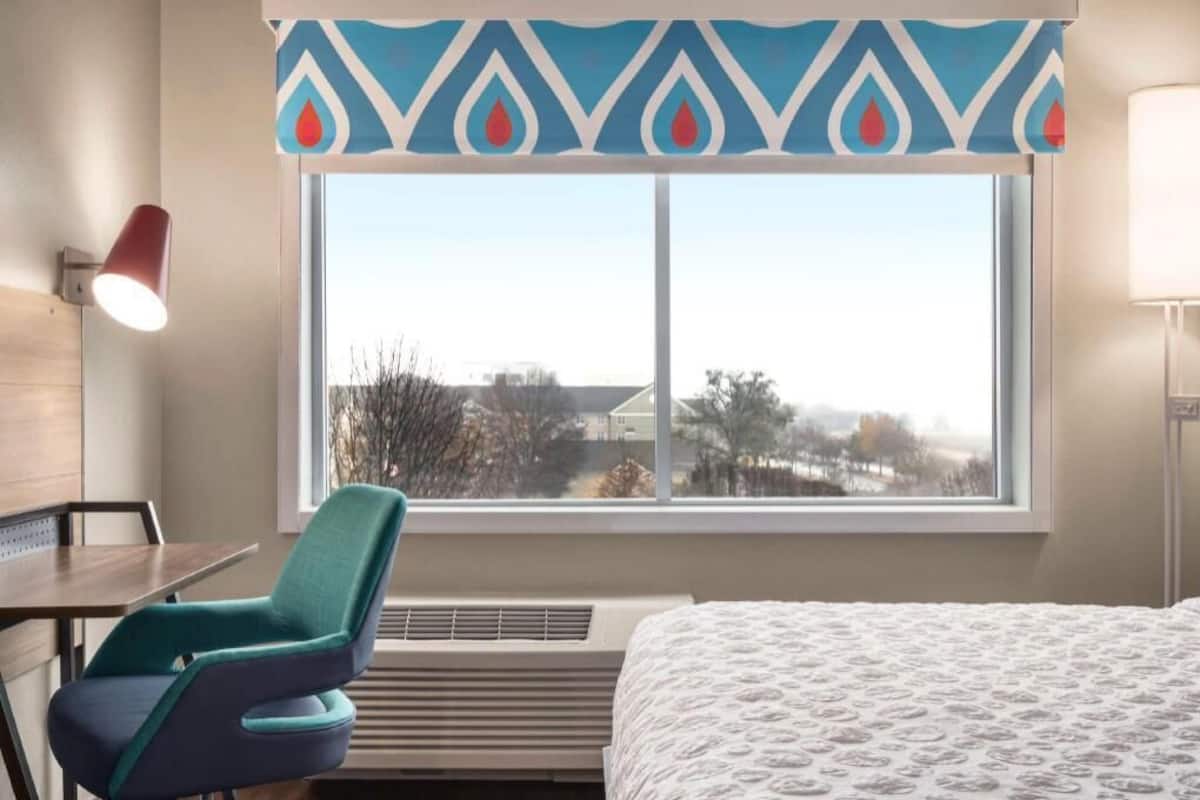 A hotel room with a bed, a desk, a chair, a window with a view, and a patterned curtain.