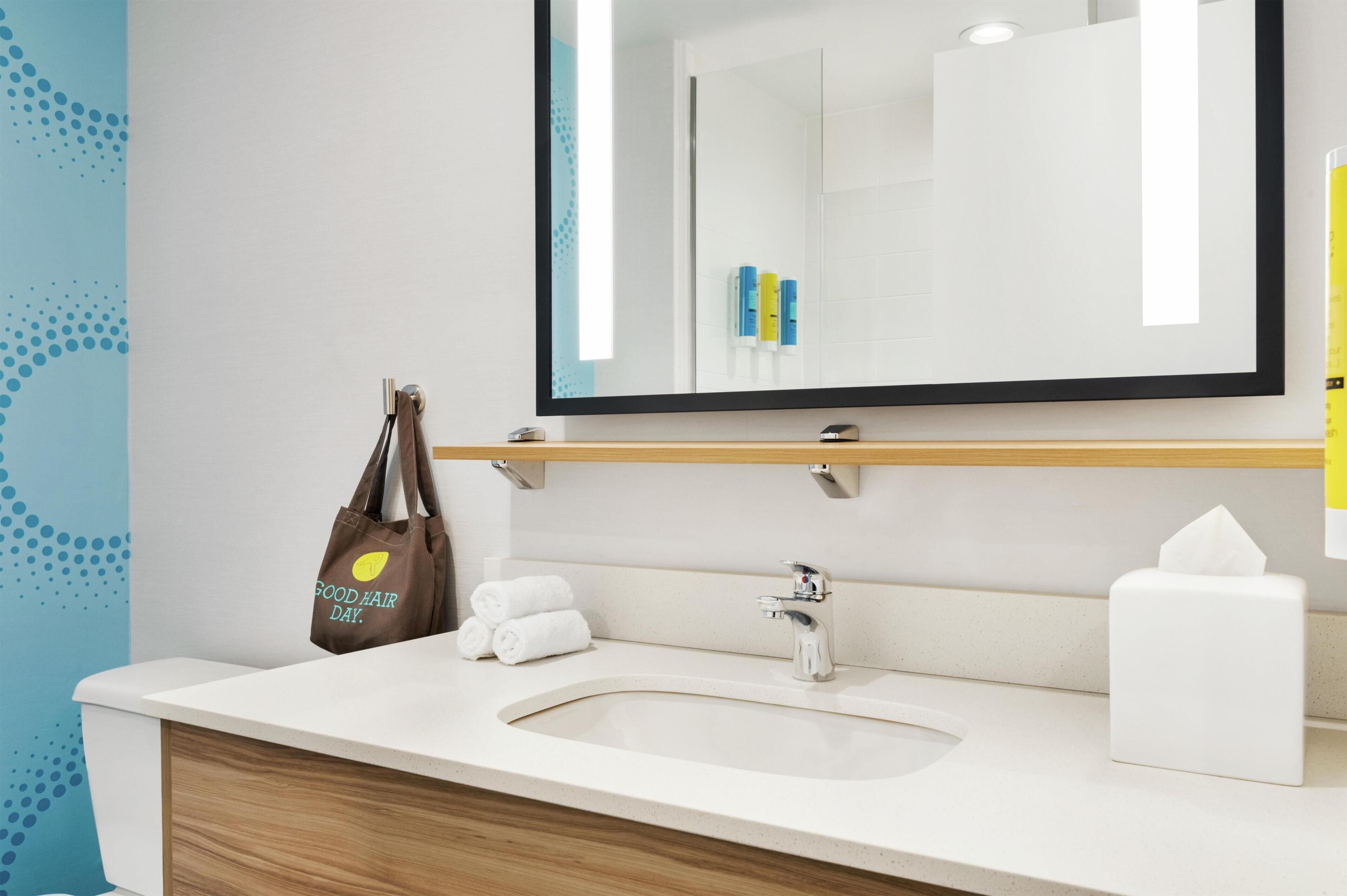 A bathroom with a white sink, a mirror, a towel rack, a tissue box, and a bag with a 'Good Hair Day' message.