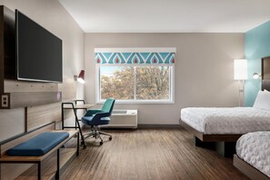 Una habitación de hotel con cama, escritorio con silla, televisor, ventana con vistas a árboles y aire acondicionado instalado en la pared.