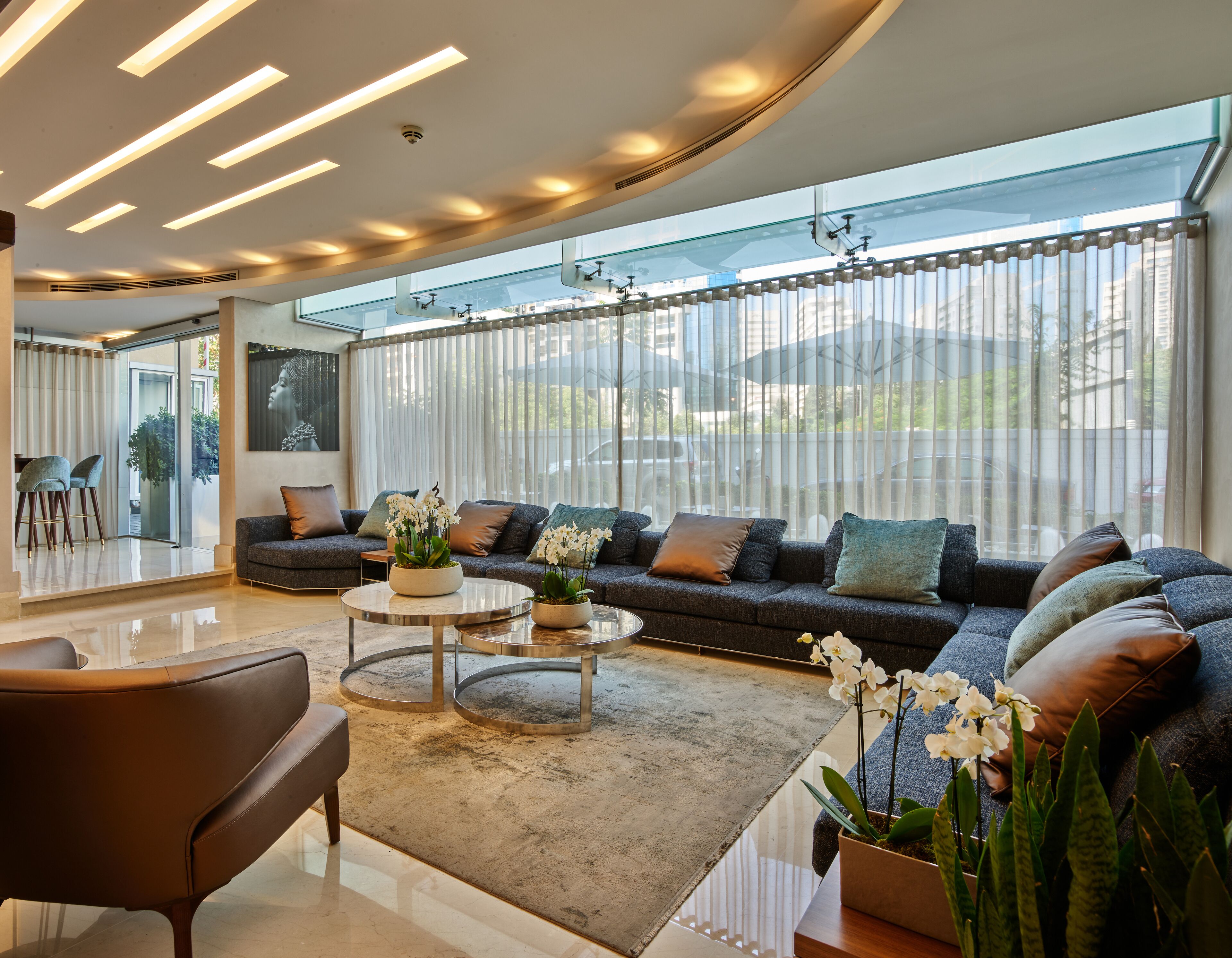 A modern living room with a sofa, coffee table, and chairs, large windows with blinds, and a ceiling with linear lighting.
