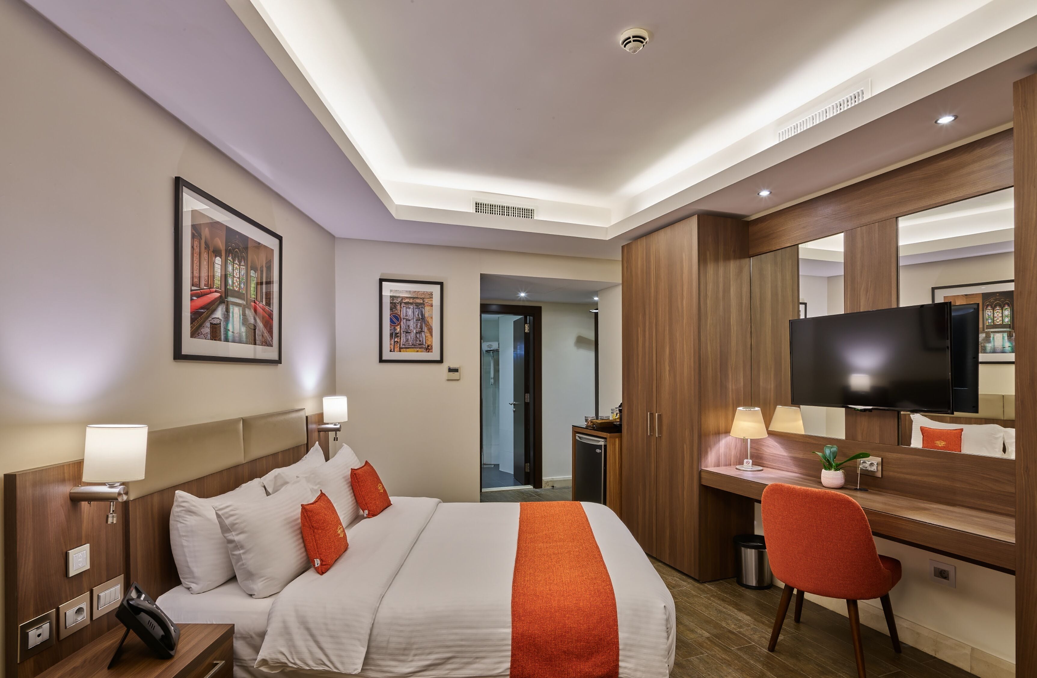 Quarto de hotel com cama, escrivaninha, cadeira, televisão e obras de arte nas paredes.