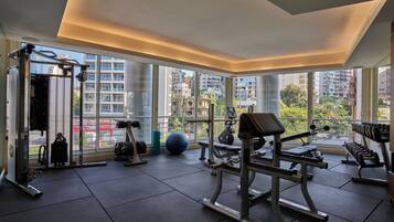 Un gimnasio con equipo de ejercicio, grandes ventanales y vistas a edificios.