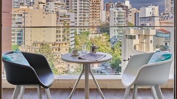 Un balcón con dos sillas y una mesita, con vista a un paisaje urbano que incluye varios edificios y un cielo nublado.