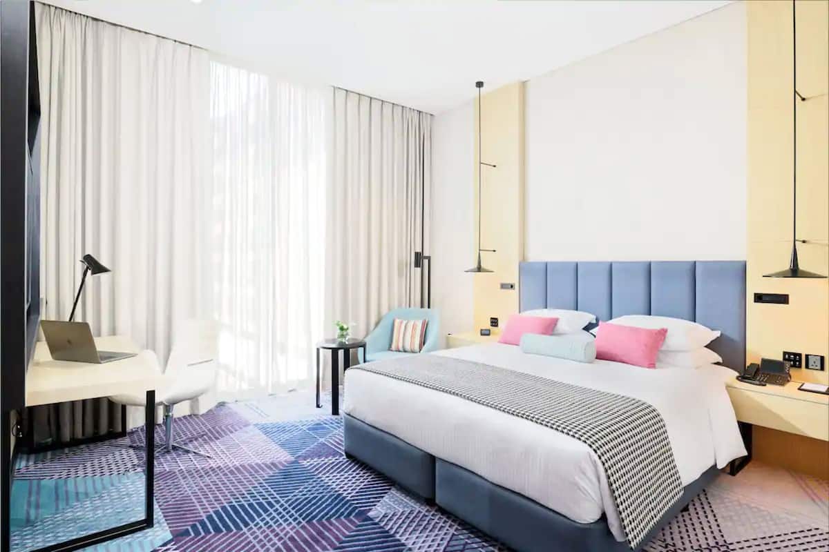 Moderni hotellihuone, jossa on suuri sänky, työpöytä kannettavalla tietokoneella, tuoli ja yöpöytä, jossa on puhelin.