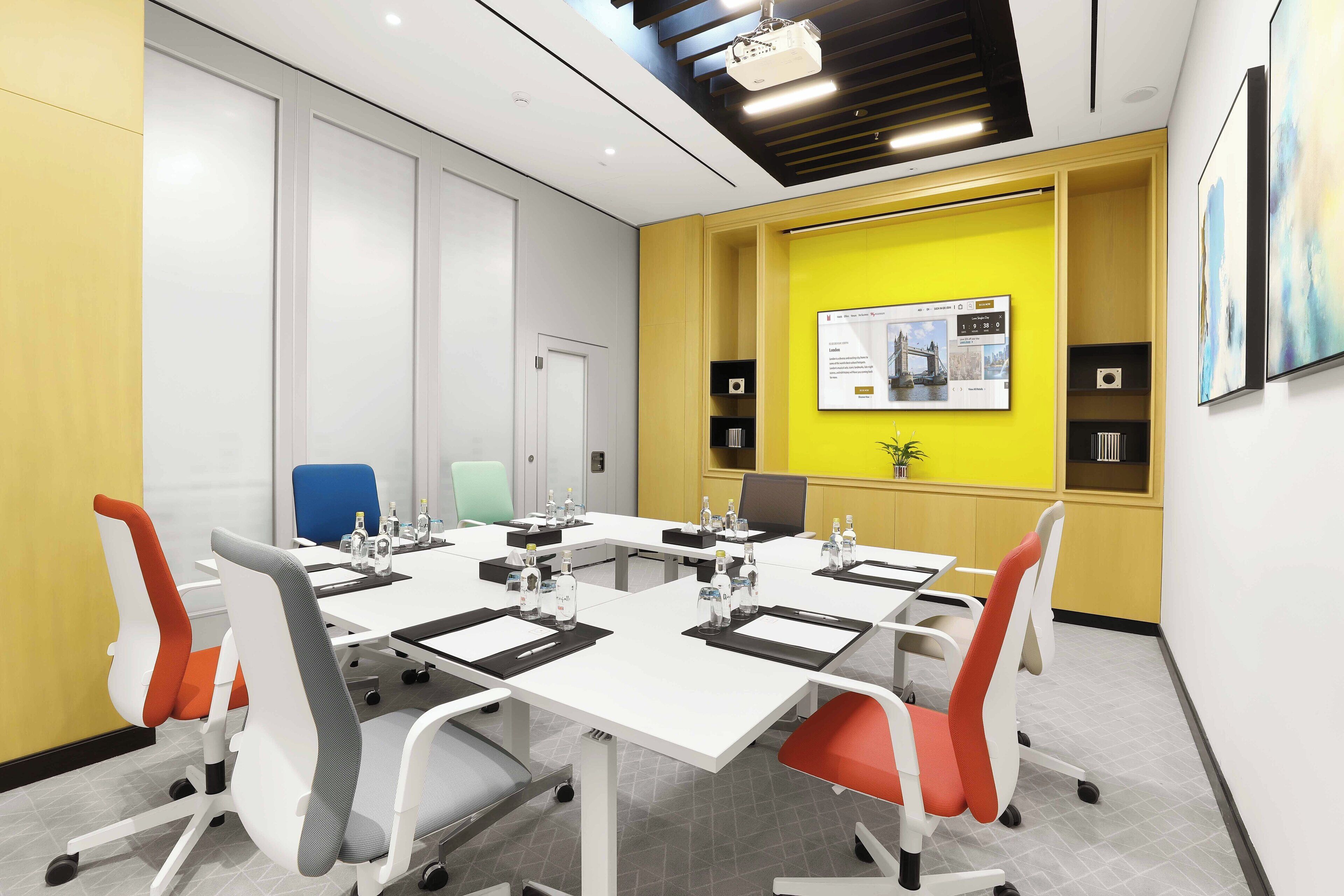 A conference room with a white table, multiple chairs, a projector, and a yellow accent wall with a framed picture.