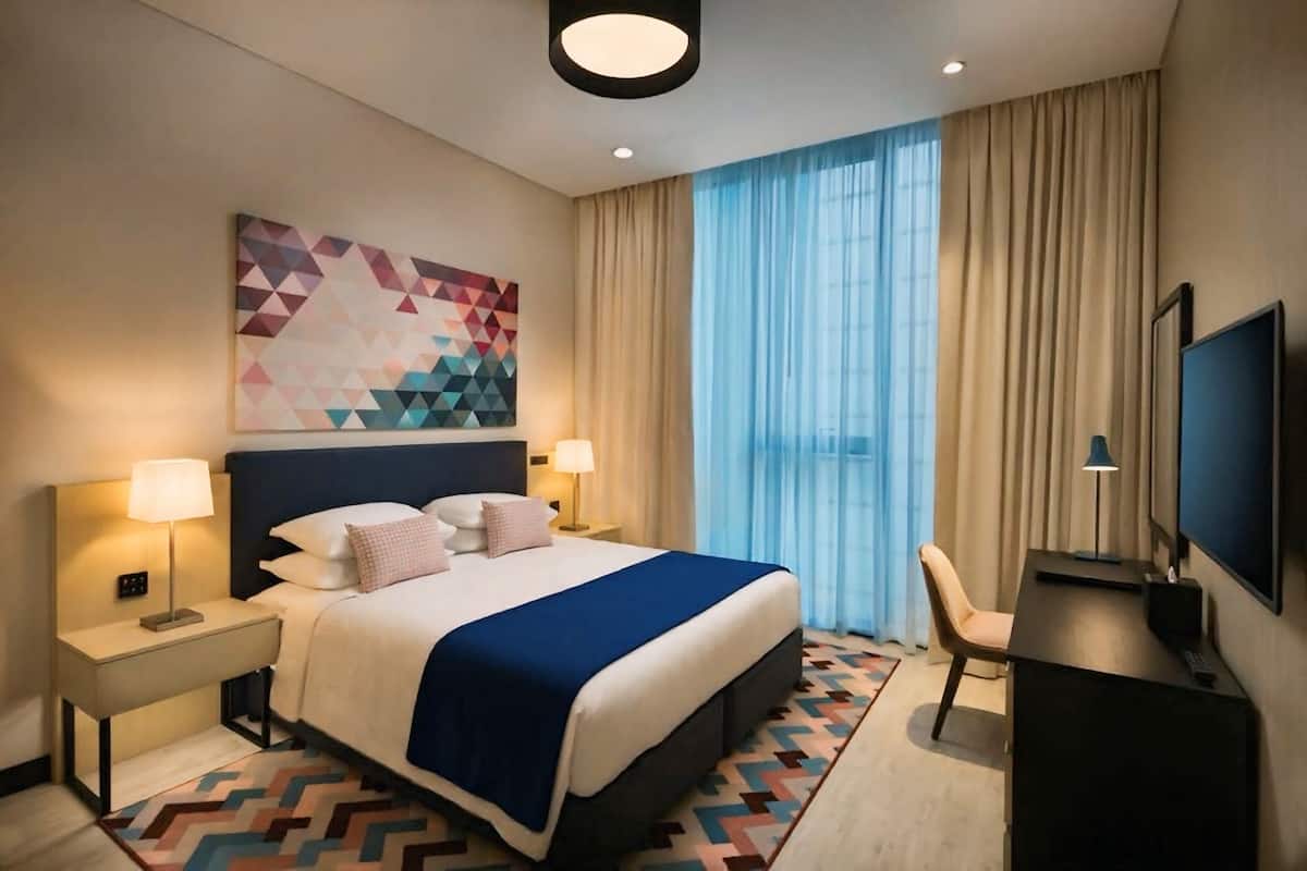 Hotellihuone, jossa on suuri sänky, kaksi yöpöytää valaisimineen, työpöytä tuolilla, taulutelevisio ja geometrisesti kuvioitu matto.